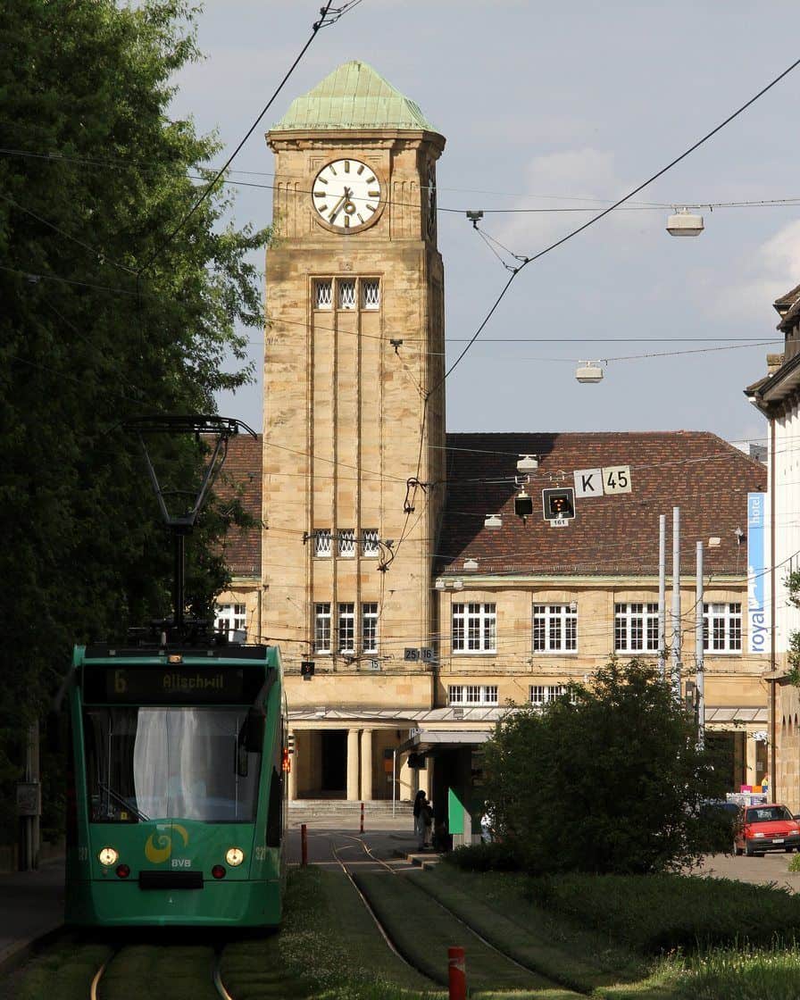 Der Badische Bahnhof in Basel, ein langgestrecktes Gebäude aus rotem Sandstein mit Uhrturm, davor ein Tram und Taxis