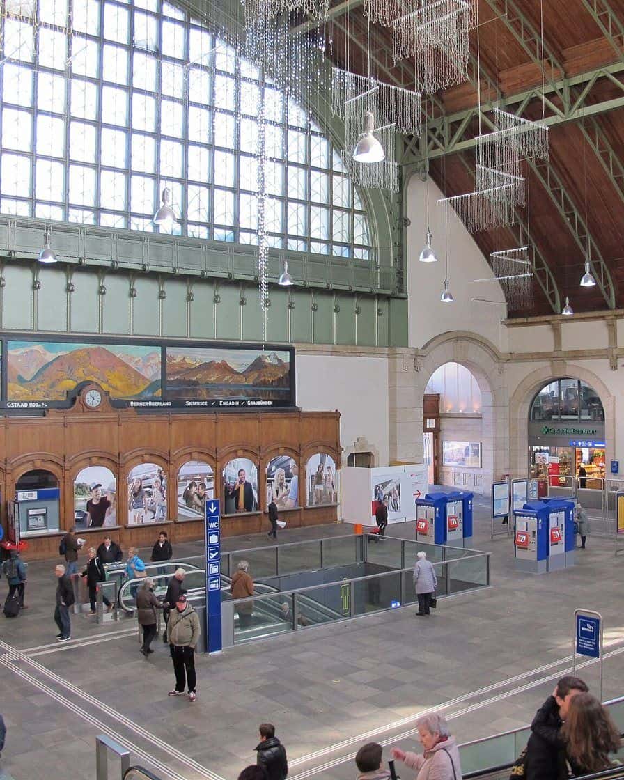 Die Schalterhalle des Bahnhofs SBB in Basel mit ihrem Glasdach, Wandbildern und Reisenden auf dem Weg zu den Gleisen