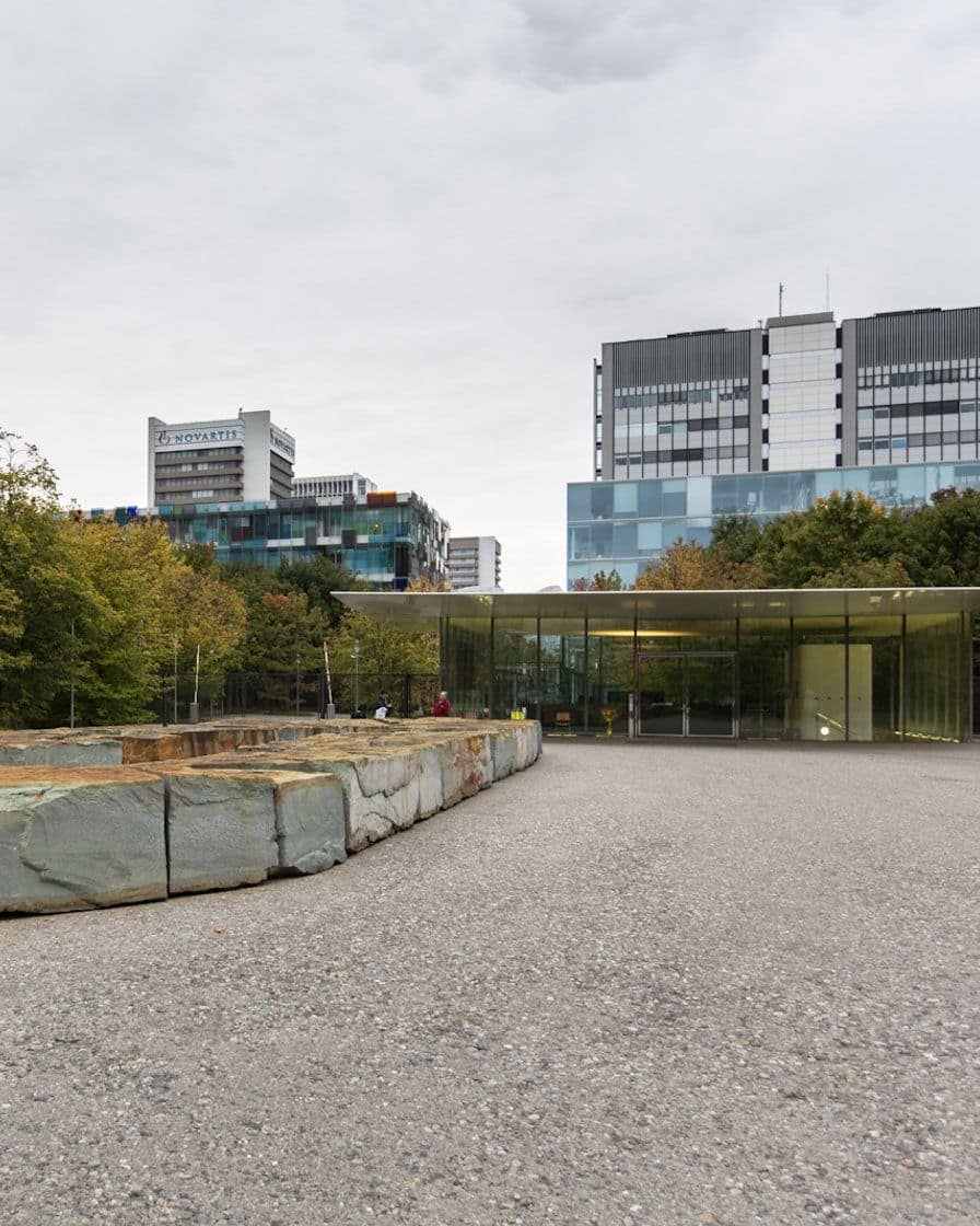 Der Novartis Campus in Basel St. Johann mit modernen Glas- und Metallfassaden an einer baumgesäumten Promenade, Passanten auf dem Weg