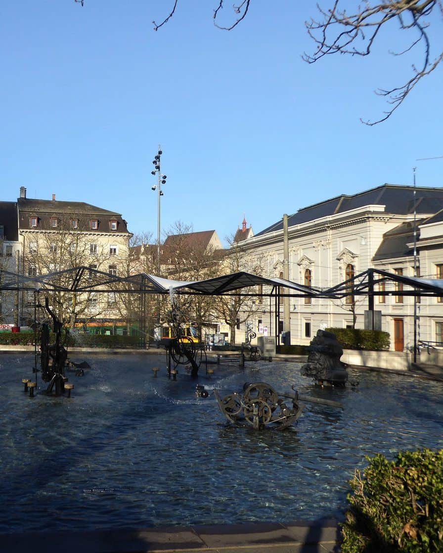 Der Tinguely-Brunnen auf dem Theaterplatz in Basel, schwarze Maschinenfiguren im flachen Becken unter Bäumen
